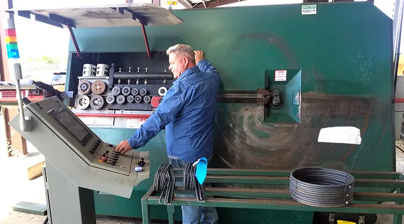 A technician at the control console of a stirrup bender, with coiled and bent rebar on the rack beside him.